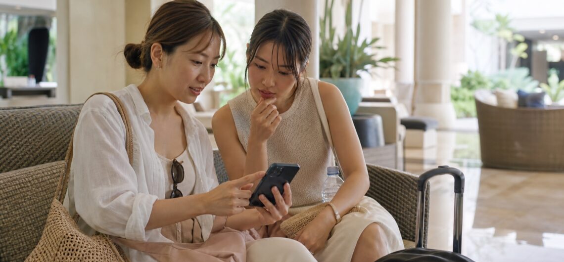 マリバゴでスマートフォンを見ながらスパを比較する日本人旅行者2人（生成画像）